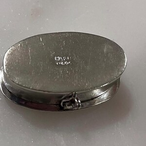 May include: Oval silver-toned hinged box with a clasp. The box is closed, showing the top and side. The top has a slightly textured surface. The word "STERLING" is stamped on the top. The box is likely for jewelry or small items.