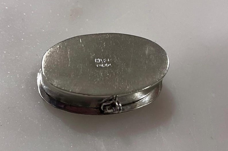 May include: Oval silver-toned hinged box with a clasp. The box is closed, showing the top and side. The top has a slightly textured surface. The word "STERLING" is stamped on the top. The box is likely for jewelry or small items.