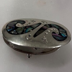 May include: Oval silver-tone trinket box with a hinged lid. The lid features an inlaid design of swirling shapes with iridescent abalone shell accents. Small star and dot details are also present on the lid. The box is closed with a small clasp.