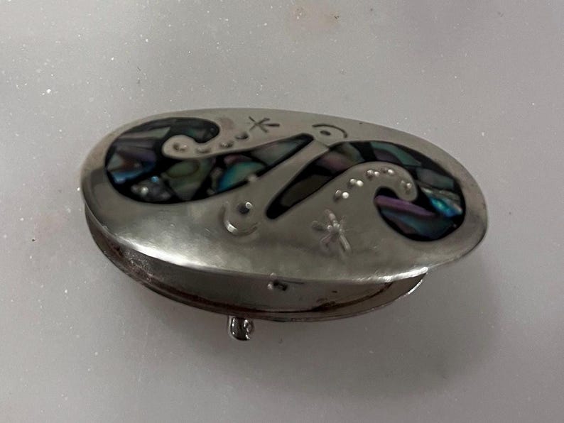 May include: Oval silver-tone trinket box with a hinged lid. The lid features an inlaid design of swirling shapes with iridescent abalone shell accents. Small star and dot details are also present on the lid. The box is closed with a small clasp.