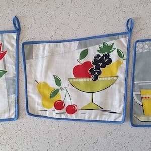 May include: Three vintage kitchen pot holders with blue trim. The first pot holder has a floral design with two red tulips in a brown pot. The second pot holder has a fruit design with a yellow pear, red cherries, and a bowl of red apples and grapes. The third pot holder has a kitchen design with a yellow cup and a white pitcher with red accents.