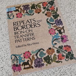 May include: A vintage book titled "Repeats and Borders Iron-On Transfer Patterns" edited by Rita Weiss. The cover features a floral border in shades of pink, blue, orange, and green. The book is on a speckled white surface.