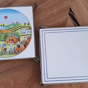 May include: Two square ceramic trivets with metal frames. One trivet features a colourful illustration of a rural scene with a hot air balloon, houses, and people. The other trivet is plain white with a blue border.