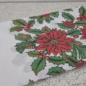 May include: White fabric with a Christmas floral pattern featuring red poinsettias, green holly leaves, and pinecones. The design is repeated across the fabric, creating a festive holiday theme.