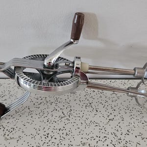 May include: Vintage hand-crank egg beater with a chrome finish and wooden handles. The beater features a gear mechanism and a rotating handle. The handle and grip are made of dark brown wood. The beater is designed for mixing ingredients.