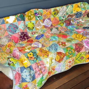 May include: A colourful patchwork quilt made from various fabrics in shades of yellow, green, pink, blue, and red. The quilt is draped over a wooden couch with a woven cane back.