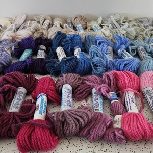 May include: A collection of yarn skeins in a variety of colors, including purple, pink, blue, and cream. The yarn is arranged in rows, with each skein displaying its color. The image is well-lit, showcasing the texture and hues of the yarn.