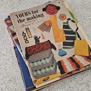May include: A pile of vintage sewing pattern books. The top book is titled "YOURS for the making" with a cartoon illustration of a girl and sewing projects. The cover features a variety of colourful sewing projects, including aprons and boxes.