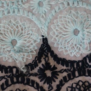 May include: A close-up of a delicate, light blue lace fabric with a floral pattern. The lace is layered over a black lace fabric with a geometric pattern.