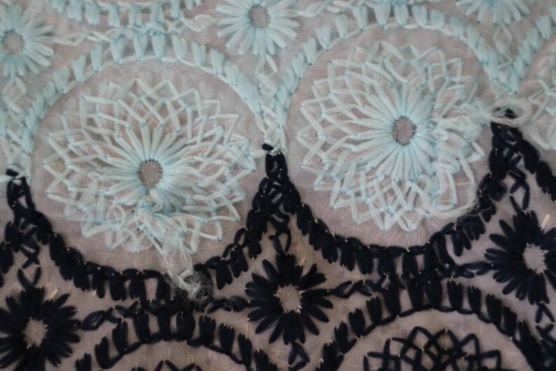 May include: A close-up of a delicate, light blue lace fabric with a floral pattern. The lace is layered over a black lace fabric with a geometric pattern.