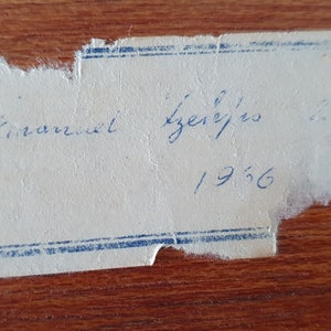 Op de afbeelding: Een wit label met blauwe rand en handgeschreven tekst die "Emmanuel Izquierdo 1956" luidt.