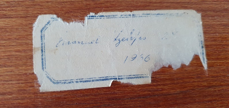 Op de afbeelding: Een wit label met blauwe rand en handgeschreven tekst die "Emmanuel Izquierdo 1956" luidt.