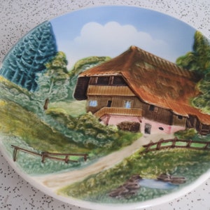 May include: A ceramic wall plate featuring a 3D design of a cottage with a thatched roof, surrounded by trees and a stream. The plate has a white background with a blue sky and white clouds.