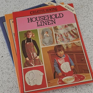 May include: A stack of vintage sewing books titled "Household Linen". The covers feature images of aprons and children's clothing. The books are in red, orange, and blue. The Singer label is visible.