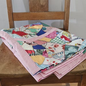May include: A folded patchwork quilt with a pink border rests on a wooden chair. The quilt displays a variety of fabric patterns in shades of blue, red, yellow, and green. The chair has a rustic, aged appearance.