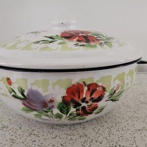 May include: White enamel bowl with lid, decorated with red, purple, and green floral patterns. The bowl has a black rim and small handles. A traditional kitchen item.