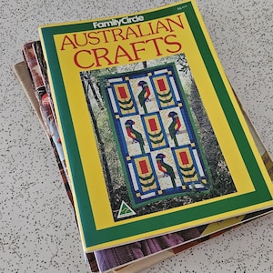 May include: A stack of vintage craft books. The top book, "Family Circle Australian Crafts," has a yellow cover with green trim and red text. The cover art features a colourful geometric design with birds. The books are on a speckled white surface.
