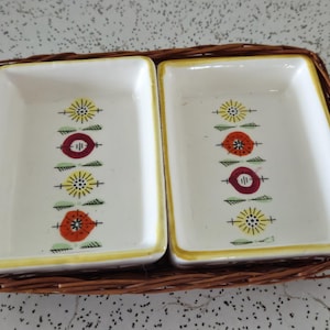 May include: A set of two white ceramic dishes with a floral design in shades of red, yellow, and green. The dishes are nestled in a woven brown basket.