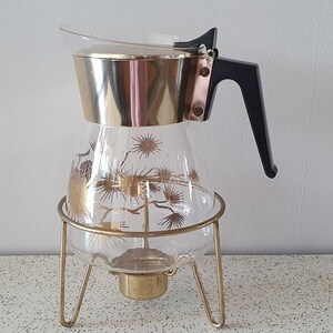 May include: A gold-toned glass coffee pot with a black handle and a gold-toned metal stand. The pot has a floral design.