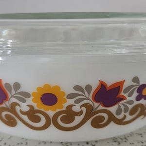 May include: White, oval casserole dish with a clear glass lid. The dish has a floral design with orange, yellow, and purple flowers, and brown scrollwork. It has two handles. A kitchenware item.