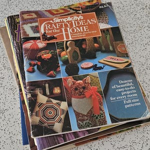 May include: A pile of vintage craft books, the top one titled "Simplicity's Crafty Ideas for the Home." The covers display colourful images of craft projects, including a star decoration, a cat, and a watermelon slice. The books are on a speckled surface.