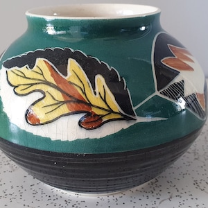 May include: A green ceramic vase with a black and white leaf design. The vase has a round body and a wide mouth.