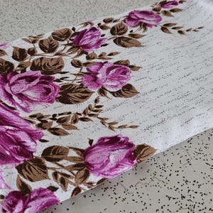 May include: White rectangular floral fabric with a pattern of large pink roses and brown leaves. The fabric has a textured background with a subtle script design. The roses are clustered along one edge.