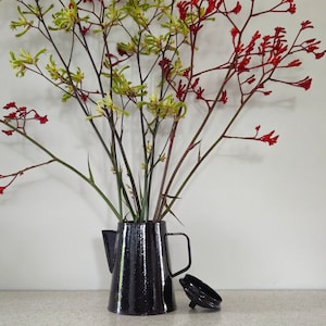 May include: A black ceramic pitcher with a speckled finish holds a bouquet of red and yellow flowers. The flowers are arranged in a vase on a table with a light-colored surface.
