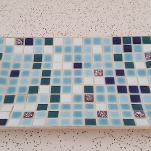 May include: A rectangular tray made of mosaic tiles in shades of blue, green, white, and purple. The tiles are arranged in a random pattern, creating a unique and eye-catching design.