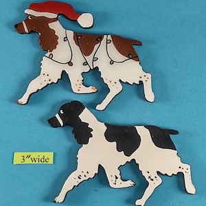 May include: Two wooden dog ornaments. One is brown and white, wearing a Santa hat and Christmas lights. The other is black and white. The ornaments are approximately 3 inches wide.