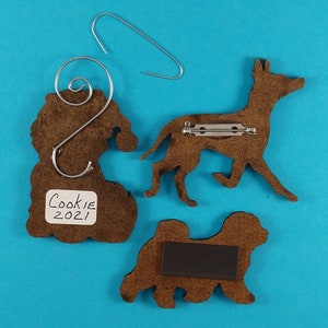May include: Three brown wooden dog ornaments with metal attachments. One ornament has a Santa hat shape with a wire hanger. Another is a dog silhouette with a pin back. The third is a dog silhouette with a magnet. The label reads "Cookie 2021".