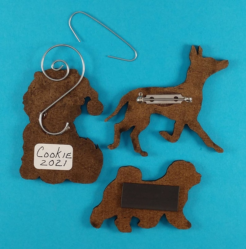 May include: Three brown wooden dog ornaments with metal attachments. One ornament has a Santa hat shape with a wire hanger. Another is a dog silhouette with a pin back. The third is a dog silhouette with a magnet. The label reads "Cookie 2021".