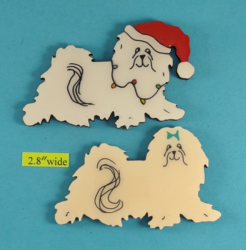 May include: Two decorative dog-shaped ornaments. One is white with a red Santa hat and string lights. The other is cream-colored with a blue bow. Both are 2.8 inches wide.