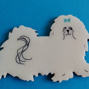 May include: A white dog-shaped ornament with black line details and a light blue bow. The dog has a fluffy appearance and a whimsical design. The background is a solid blue color.
