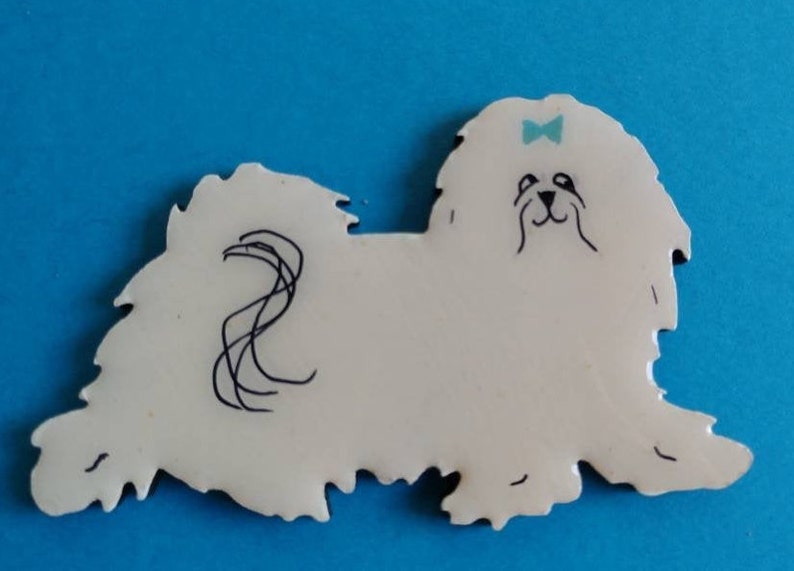 May include: A white dog-shaped ornament with black line details and a light blue bow. The dog has a fluffy appearance and a whimsical design. The background is a solid blue color.