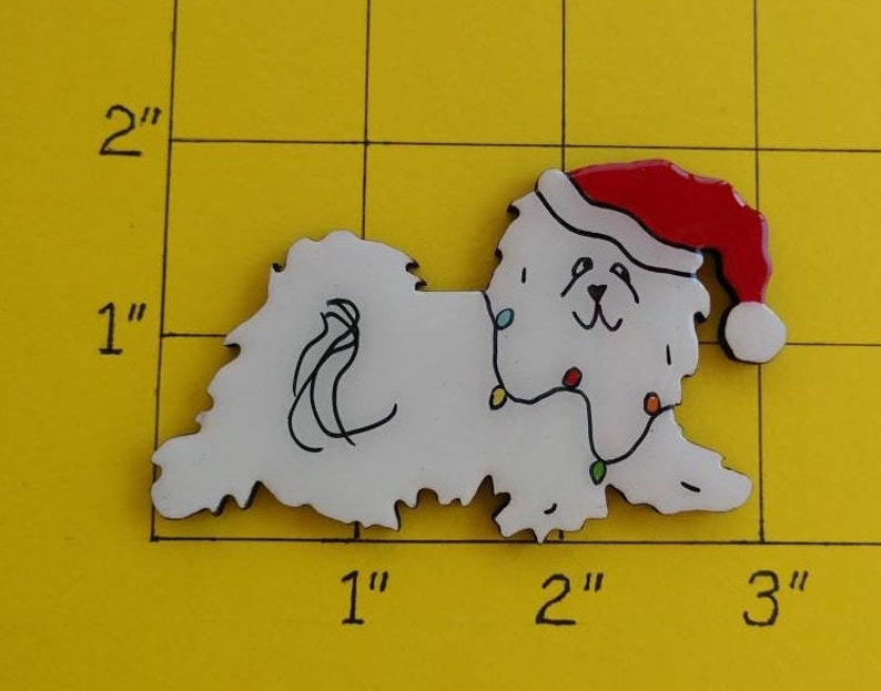 May include: A white dog-shaped Christmas ornament, approximately 2 inches long, wearing a red Santa hat and a string of colorful lights. The ornament has black details and is on a yellow grid background.