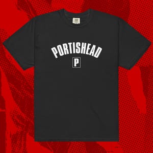 May include: Black t-shirt with the word "PORTISHEAD" in white, arched across the chest. Below the word is a white square with the letter "P" inside. The shirt is a crew neck style.