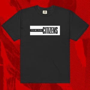 May include: Black short-sleeved t-shirt with the text "THE N.Y. CITIZENS" in a white rectangular box. The shirt has a crew neck and a tag at the back of the neck. The background is a red and black halftone pattern.