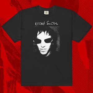 Può includere: T-shirt nera con un'immagine in bianco e nero di una persona con occhiali da sole e il nome "Elliott Smith" in scrittura bianca. La maglietta è realizzata in un materiale morbido.