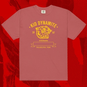 May include: A dusty rose t-shirt with a yellow graphic that reads "KID DYNAMITE" above a roaring cougar head. Below the graphic is a banner with "COUGARS" and "PHILADELPHIA, PENN." printed on it. The shirt has a crew neck and short sleeves.