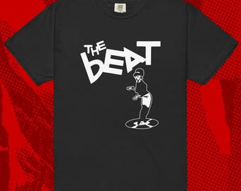 Puede incluir: Camiseta negra con la inscripción blanca "THE BEAT" y una ilustración blanca de una persona sobre un disco. La camiseta tiene cuello redondo y mangas cortas. El diseño está centrado.