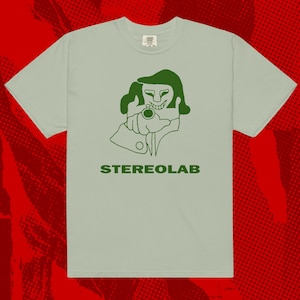 Puede incluir: Camiseta verde claro con una ilustración verde de una persona apuntando con una pistola. La palabra "STEREOAB" está impresa en verde debajo. La camiseta tiene mangas cortas y cuello redondo.