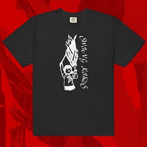 May include: Black t-shirt with a white graphic of a child holding a flag and the text "SWANG KAOS" in a vertical font. The design is a simple, illustrative style. The shirt has short sleeves and a crew neck.