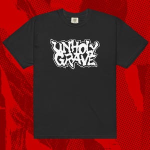 May include: Black t-shirt with the white text "UNHOLY GRAVE" in a distressed, gothic font. The shirt has short sleeves and a crew neck. The background is a red and black pattern.