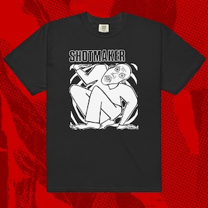 May include: Black t-shirt with a white graphic of a person with a shocked expression, surrounded by a swirling pattern. The text "SHOTMAKER" is printed above the graphic.