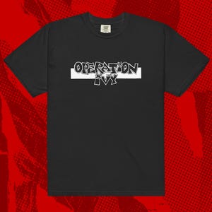 Puede incluir: Camiseta negra con el texto blanco "OPERATION IVY" en estilo graffiti. El texto está centrado en una barra horizontal blanca. La camiseta tiene mangas cortas y cuello redondo.