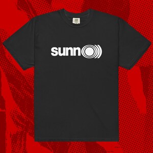 May include: Black t-shirt with the word "sunn" in white, accompanied by a white circular design. The shirt has short sleeves and a crew neck. The background is red with a halftone pattern.