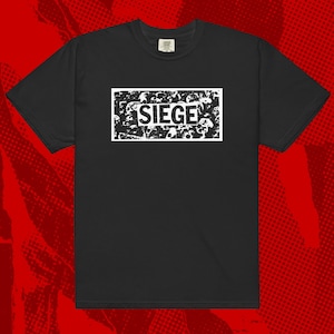 Puede incluir: Camiseta negra con un gráfico rectangular blanco. El gráfico presenta la palabra "SIEGE" en negrita y mayúsculas, sobre un fondo texturizado. La camiseta tiene mangas cortas y cuello redondo.