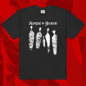 May include: A black short-sleeve t-shirt featuring a white graphic print on the chest. The design displays the text "REASON TO BELIEVE" in a stylized font above four abstract, textured white vertical shapes resembling silhouettes of figures.