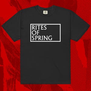 Puede incluir: Camiseta negra con el texto blanco "RITES OF SPRING" dentro de un marco rectangular blanco. La camiseta tiene mangas cortas y cuello redondo. El fondo es un patrón de semitonos rojo.